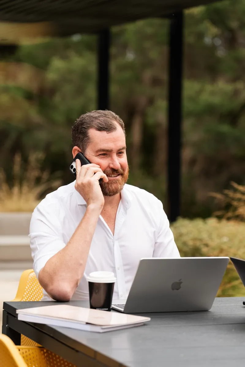A Future Finance Group broker smiling on a phone call at an outdoor table with a laptop and coffee