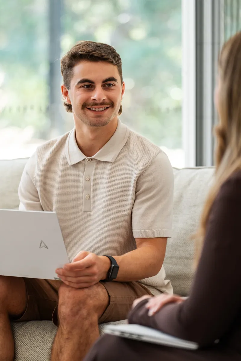 A smiling young man in a knit polo holding a laptop on a sofa during a meeting